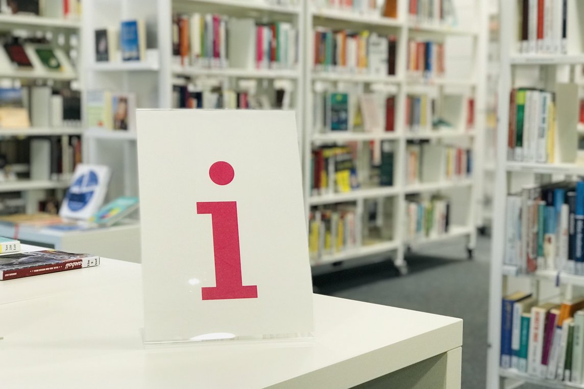Auf dem Foto ist ein Aufsteller mit dem Buchstaben i in pink zu sehen, auf einem weißen Tisch. Im Hintergrund sind die Bücherregale der Stadtbücherei zu sehen.