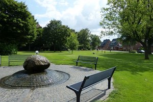 Das Bild zeigt den Von-Galen-Park in Merfeld mit Bänken und einem Wasserspiel im Vordergrund.