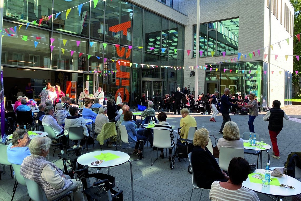 Das Bild zeigt Menschen an Bistro-Tischen, tanzende paar und ein Orchester auf dem Markt der Möglichkeiten.